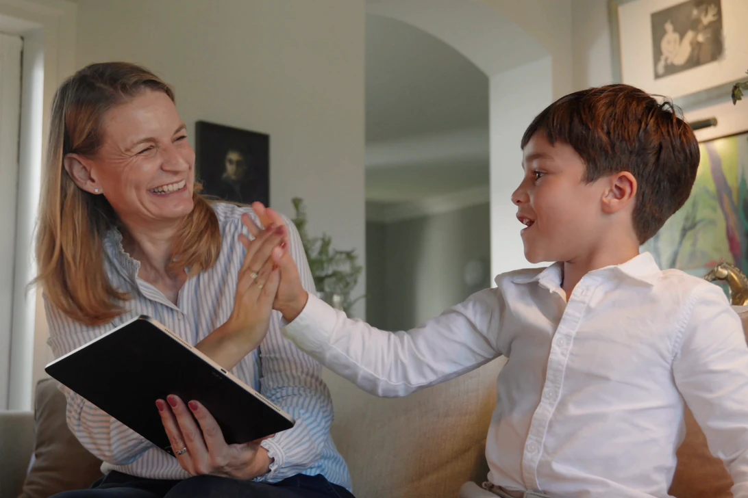 A parent and a child laugh and high-five on a sofa, a tablet in the parent's hand